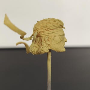 Peut inclure: Une sculpture de t&ecirc;te miniature en r&eacute;sine non peinte de couleur beige repr&eacute;sentant un personnage aux cheveux courts textur&eacute;s, portant un bandeau nou&eacute; &agrave; l'arri&egrave;re avec deux rubans flottants et un cache-&oelig;il, fix&eacute;e sur une fine tige verticale.