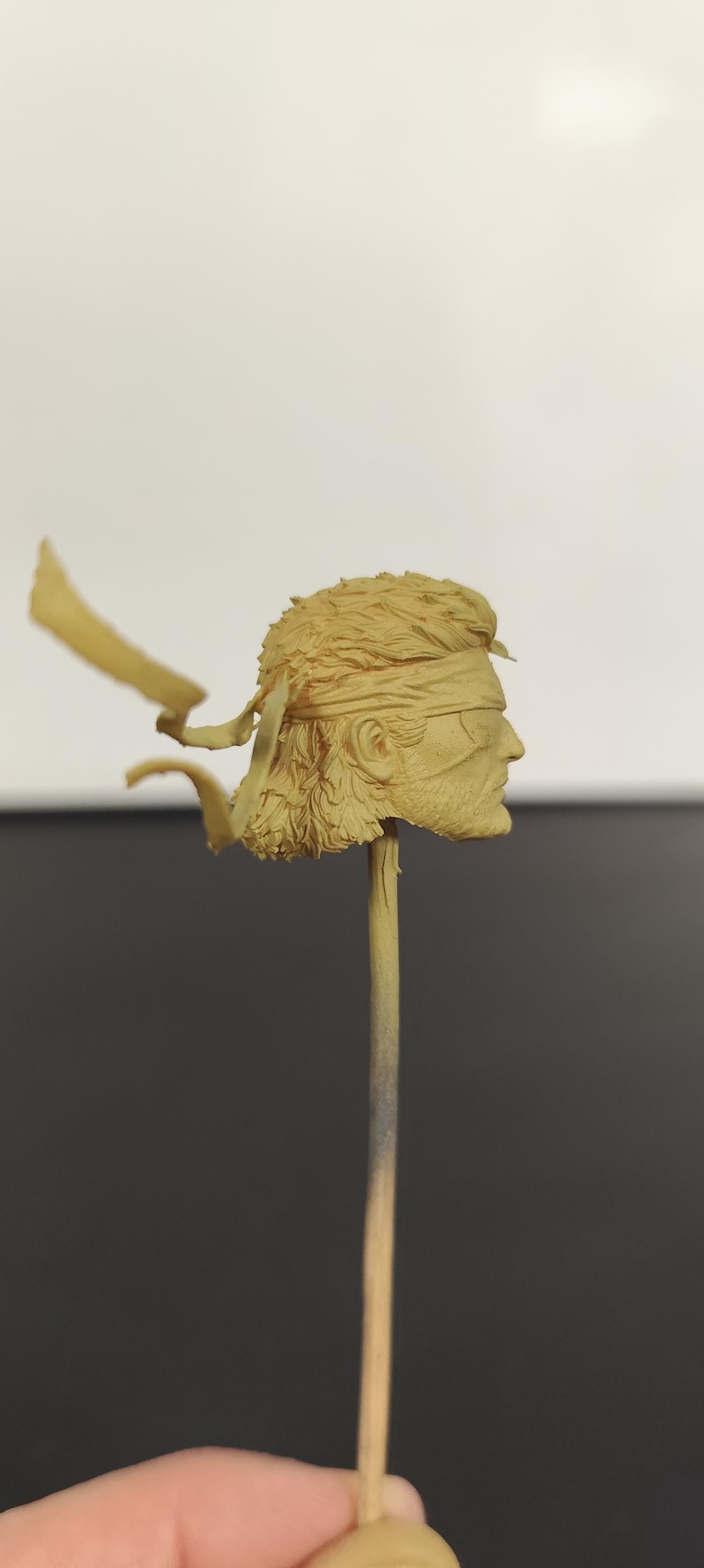 Peut inclure: Une sculpture de t&ecirc;te miniature en r&eacute;sine non peinte de couleur beige repr&eacute;sentant un personnage aux cheveux courts textur&eacute;s, portant un bandeau nou&eacute; &agrave; l'arri&egrave;re avec deux rubans flottants et un cache-&oelig;il, fix&eacute;e sur une fine tige verticale.
