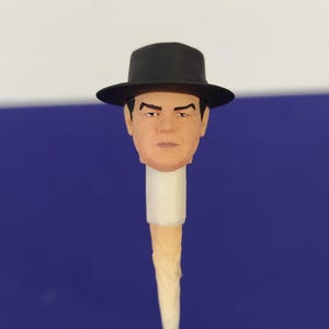 Peut inclure: Une tête sculptée miniature d'une personne au teint clair, aux sourcils peints en foncé et à l'expression neutre. La tête porte un chapeau fedora noir et est montée sur un support cylindrique blanc fixé à un fin bâton en bois.