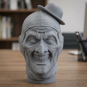 Puede incluir: Un busto escultórico gris monocromático de una cabeza de payaso con una amplia sonrisa, ojos hundidos y un pequeño sombrero de bombín posado en un lado de la cabeza. La figura tiene un acabado mate liso con arrugas y texturas faciales detalladas.