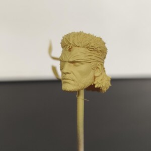 Peut inclure: Une sculpture en r&eacute;sine non peinte de couleur beige repr&eacute;sentant une t&ecirc;te humaine avec une coiffure mulet textur&eacute;e, un bandeau et des traits du visage d&eacute;taill&eacute;s, incluant une barbe et une moustache. La t&ecirc;te est fix&eacute;e sur une fine tige verticale.