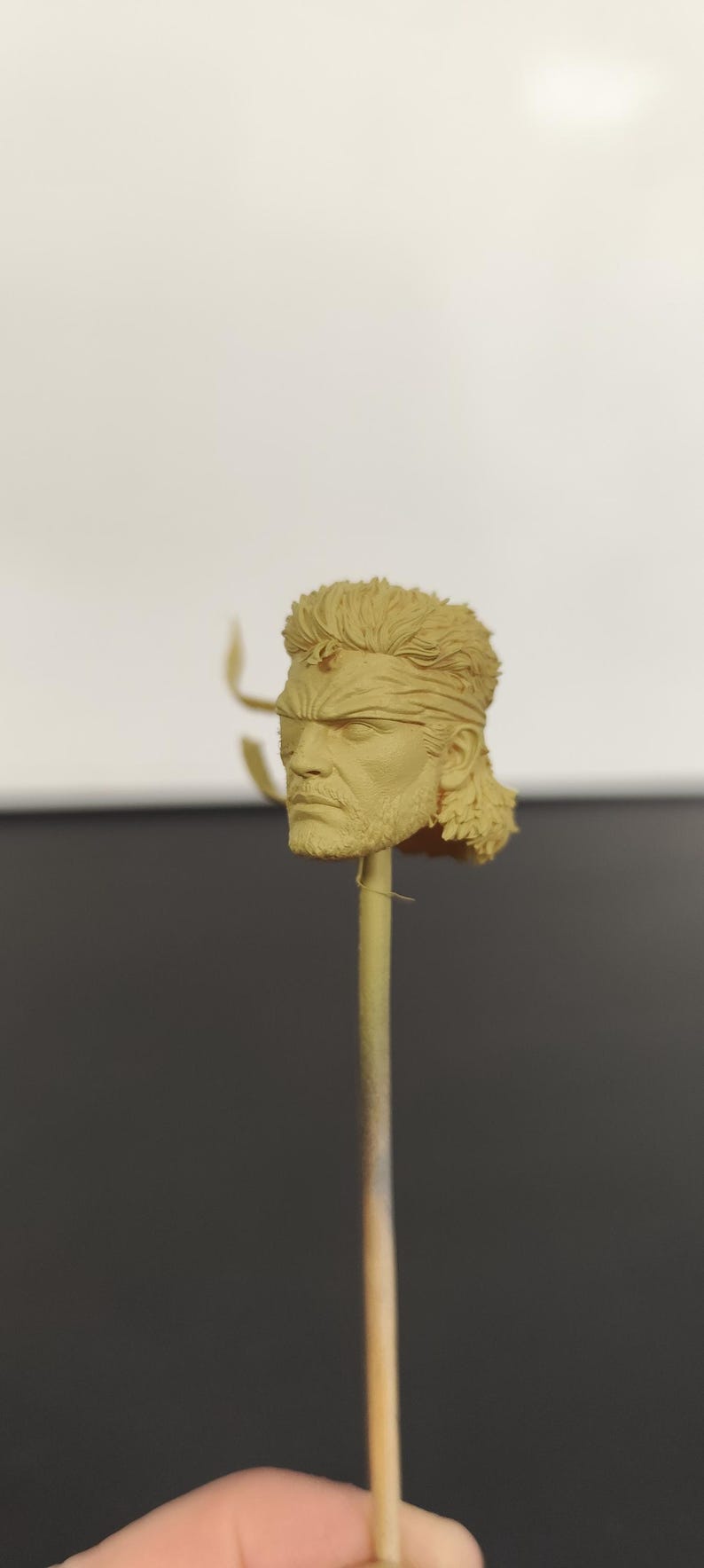 Peut inclure: Une sculpture en r&eacute;sine non peinte de couleur beige repr&eacute;sentant une t&ecirc;te humaine avec une coiffure mulet textur&eacute;e, un bandeau et des traits du visage d&eacute;taill&eacute;s, incluant une barbe et une moustache. La t&ecirc;te est fix&eacute;e sur une fine tige verticale.