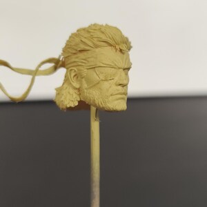Peut inclure: Une sculpture de t&ecirc;te en r&eacute;sine non peinte de couleur beige repr&eacute;sentant un personnage avec une coiffure courte et textur&eacute;e, une barbe prononc&eacute;e et un cache-&oelig;il sur l'&oelig;il gauche. La t&ecirc;te est fix&eacute;e sur une fine tige verticale pour la peinture.
