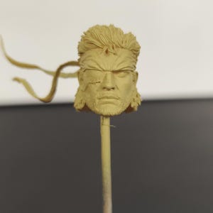 Peut inclure: Une t&ecirc;te sculpt&eacute;e monochrome de couleur beige repr&eacute;sentant un personnage aux cheveux courts et textur&eacute;s, avec un front marqu&eacute; et un cache-&oelig;il distinct sur l'&oelig;il droit. Le visage affiche une expression s&eacute;rieuse avec une barbe et un bandeau fin.