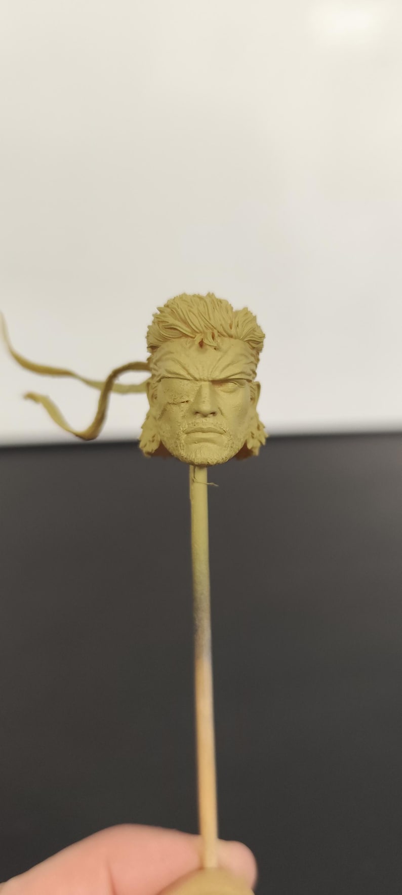 Peut inclure: Une t&ecirc;te sculpt&eacute;e monochrome de couleur beige repr&eacute;sentant un personnage aux cheveux courts et textur&eacute;s, avec un front marqu&eacute; et un cache-&oelig;il distinct sur l'&oelig;il droit. Le visage affiche une expression s&eacute;rieuse avec une barbe et un bandeau fin.