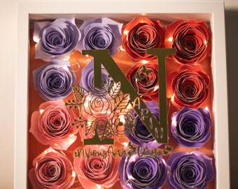 May include: A white frame with a glass front containing a gold letter N with floral accents and a string of lights. The frame is filled with paper flowers in shades of purple, pink, and orange.