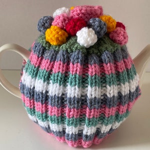 May include: A cream-coloured teapot with a knitted tea cosy. The cosy has horizontal stripes in pink, white, grey, and green. The top of the cosy is decorated with multi-coloured yarn pom-poms. A charming, handmade item.