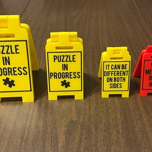 May include: Four yellow and red construction style signs with black text. The signs read "Puzzle in Progress", "Puzzle in Progress", "It can be different on both sides", and "Your message right here".