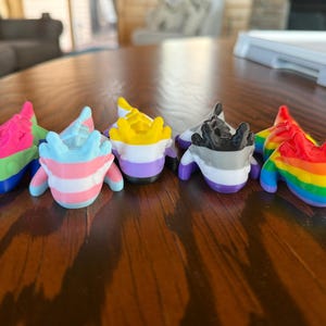 Peut inclure: Six petites figurines colorées d'axolotls, chacune peinte avec un motif de drapeau de la fierté différent. Les axolotls sont disposés en rangée de gauche à droite : transgenre, non binaire, bisexuel, asexuel, gris asexuel et arc-en-ciel.