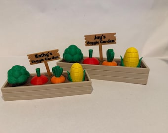 May include: Two miniature wooden garden boxes with signs that read "Kathy's Veggie Garden" and "Jay's Veggie Garden". Each box contains a variety of colorful vegetables, including broccoli, carrots, corn, and radishes.