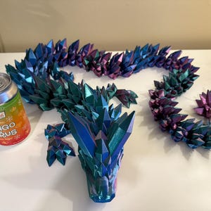 May include: A 3D-printed dragon sculpture in shades of blue, green, and purple, with a crystalline texture. The dragon is segmented, with a head, body, and tail. A can of Mango Citrus soda is next to the dragon.