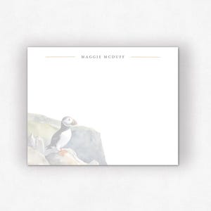 May include: A white stationery card with a watercolor illustration of a puffin bird perched on a rock. The card has the text "MAGGIE MCDUFF" in a simple font, centered at the top. The overall design is soft and muted.