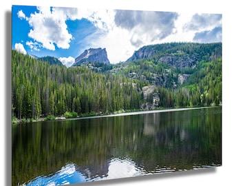 May include: A scenic landscape photograph featuring a mountain lake with a reflection of the surrounding forest and a mountain peak in the distance. The sky is blue with white clouds.