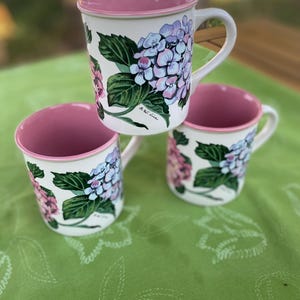 May include: Three ceramic mugs with a white exterior and a pink interior. Each mug features a painted design of blue and pink hydrangea flowers with green leaves. The signature S.W. Lee is visible on the side of the mugs near the floral pattern.