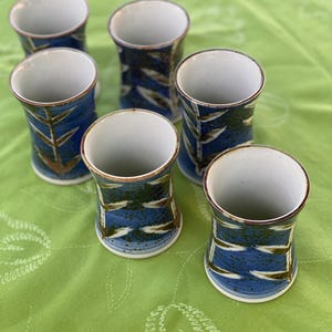 May include: A set of six ceramic tumblers with a white interior and a blue exterior featuring a speckled texture and a repeating brown and white leaf-like pattern. Each cylindrical cup has a slightly flared rim and a smooth, glazed finish.