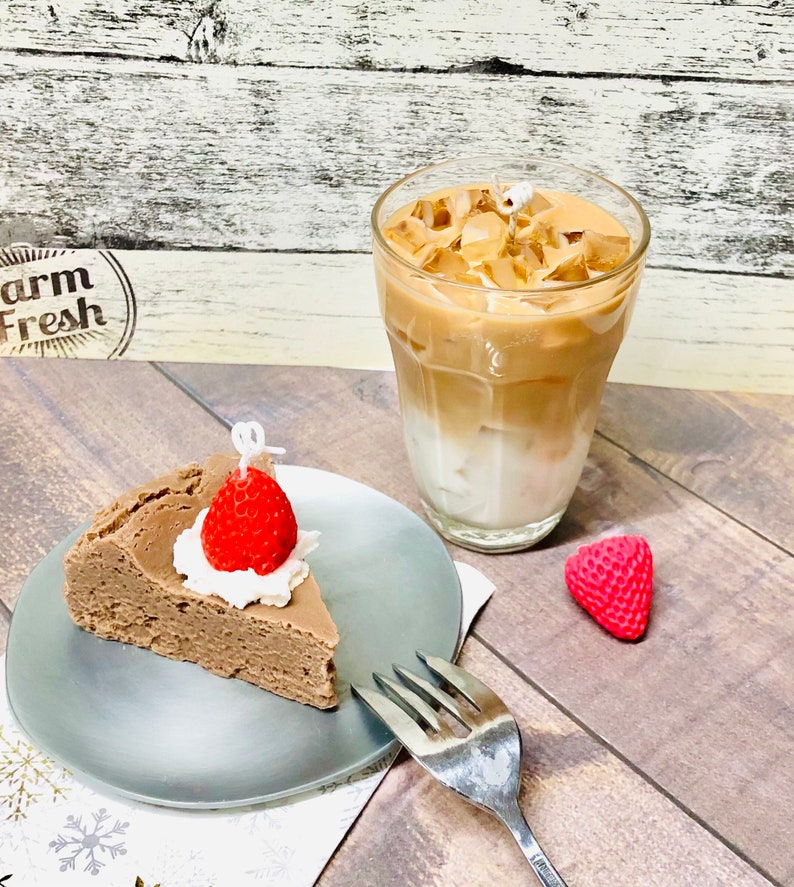 May include: A slice of chocolate cake with whipped cream and a strawberry on top, served on a silver plate with a fork. A glass of iced coffee is in the background.