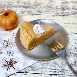 May include: A slice of pumpkin cheesecake with whipped cream on top, on a silver plate with a fork beside it.