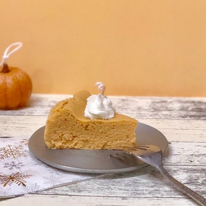 May include: A slice of pumpkin cheesecake topped with whipped cream on a silver plate with a fork. The cheesecake is on a white and gold snowflake patterned napkin.