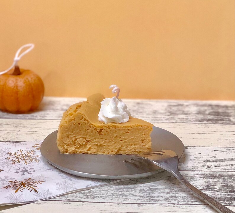 May include: A slice of pumpkin cheesecake topped with whipped cream on a silver plate with a fork. The cheesecake is on a white and gold snowflake patterned napkin.