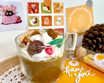 Könnte beinhalten: Eine Kerze in einem klaren Glasbecher mit brauner Basis, weißer Schlagsahne und dekorativen Toppings wie Keksen und Süßigkeiten. Eine Karte mit "thank you" ist sichtbar.