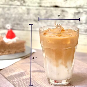 May include: A clear glass with a 3 inch diameter and 4.5 inch height filled with iced coffee. The coffee is layered with a light brown layer on top of a white layer.