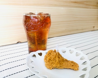 May include: A glass of cola with ice cubes and a fried chicken leg candle on a white plate with a heart-shaped design.