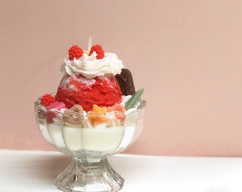May include: A red and white ice cream sundae candle in a clear glass dish. The candle is decorated with white whipped cream, red and white sprinkles, and a green leaf.