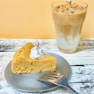 May include: A slice of fluffy cheesecake with whipped cream on top, served on a silver plate with a fork. A glass of iced coffee with ice cubes is in the background.