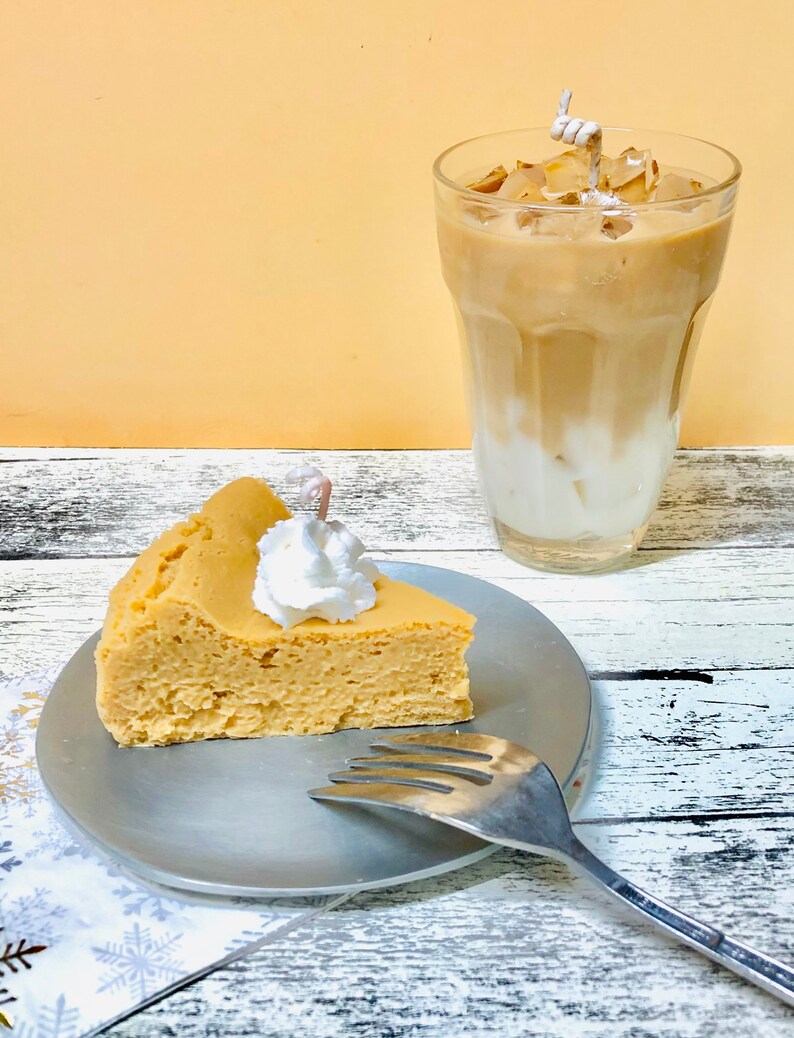 May include: A slice of fluffy cheesecake with whipped cream on top, served on a silver plate with a fork. A glass of iced coffee with ice cubes is in the background.
