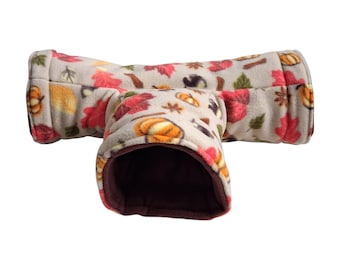 May include: A T-shaped pet tunnel made of soft beige fleece fabric. The exterior features a repeating pattern of orange pumpkins, red and green autumn leaves, and brown star anise. The interior of the tunnel is lined with solid dark brown fabric.