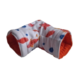 Puede incluir: Un túnel para mascotas en forma de L hecho de tela polar blanca suave con un patrón repetitivo de peces naranjas, estrellas de mar rojas y pequeños puntos azules. El interior del túnel está forrado con una tela polar de color naranja sólido.