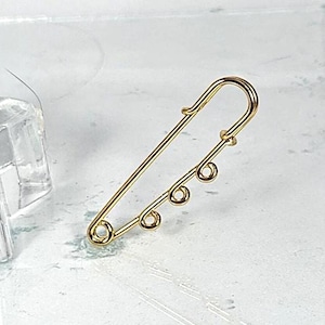 May include: A gold-colored safety pin with three small decorative loops. The pin is a classic design, with a curved top and a pointed end. The pin is likely made of metal and is shown against a light background.