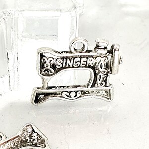 以下が含まれることがあります： 前面に「SINGER」の文字が入った、銀色のミシンチャーム。チャームには複雑なディテールがあり、ネックレスやブレスレットに取り付けるためのループが上部にあります。チャームは、ヴィンテージミシンのミニチュアレプリカです。
