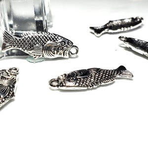Puede incluir: Una colección de dijes de metal plateado con forma de pez, cada uno con texturas de escamas detalladas, aletas y un pequeño lazo circular en la cabeza para su fijación. Los dijes están dispuestos en varias orientaciones, mostrando el diseño en relieve.