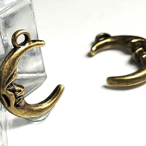 May include: Two antique gold-colored crescent moon charms with a face carved into the moon's surface. One charm is attached to a clear glass object. The charms are on a white surface.
