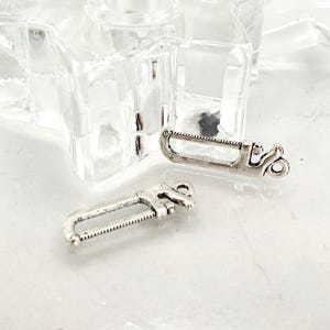 May include: Two silver-colored saw charms. Each charm has a U-shaped frame with a toothed blade and a decorative handle. The charms are small and detailed, suitable for jewelry making or crafting projects.