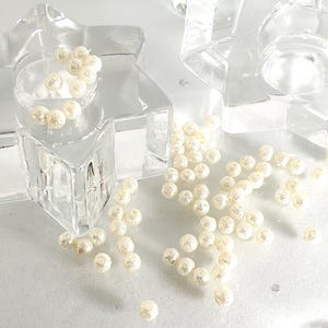 Könnte beinhalten: Klare, sternförmige Kerzenhalter aus Glas mit verstreuten elfenbeinfarbenen Perlen. Die Kerzenhalter haben ein geometrisches Design, und die Perlen sind in der Mitte und darum herum verstreut.