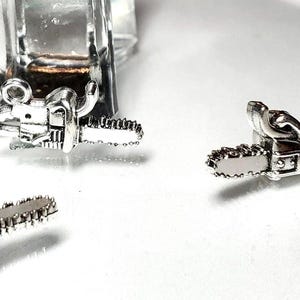 May include: Close-up of several small, silver-colored chainsaw charms. The charms have detailed saw blades and handles, and are likely used for jewelry making or crafting. The background is a bright white surface.