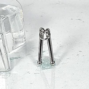 May include: A small, silver-colored metal tool with a looped top and two legs that end in small, round balls. The tool is set against a white and gray background.