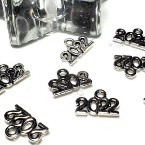 May include: Silver charms with the year 2022 printed on them. The charms are scattered on a white surface.