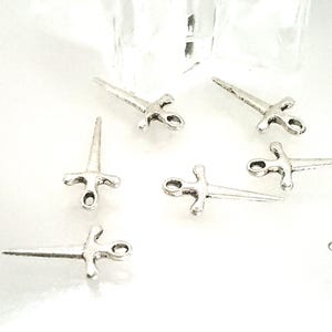 May include: Small, silver-colored sword charms with a cross-shaped handle and a pointed blade. These charms are scattered across a white surface, suggesting a craft or jewelry-making theme.