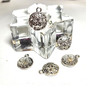 May include: Silver-toned sand dollar charms with a detailed starfish design. These small, round charms have a loop at the top for easy attachment to jewelry or other crafts. The charms are displayed on a clear, star-shaped glass object.