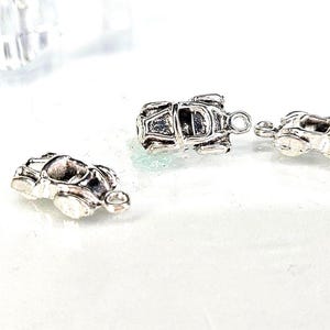 May include: Three small, silver-colored car charms. Each charm has a detailed design of a classic car, with visible headlights, wheels, and a rounded body. The charms are likely for jewelry making or crafting.