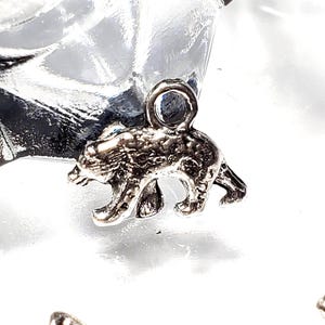May include: A small, silver-colored bear charm with a loop at the top for attaching to jewelry or other crafts. The bear is depicted in a walking pose, with detailed fur texture. The charm is likely made of metal.