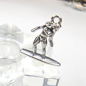 May include: A silver-colored charm depicting a surfer riding a surfboard. The surfer is in a dynamic pose, with arms outstretched. The charm has a small loop at the top for attaching to a bracelet or necklace.