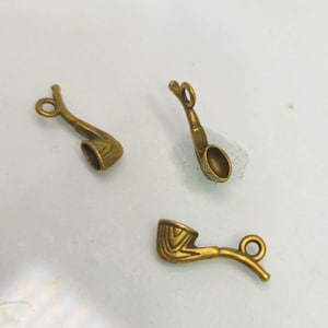 May include: Three small, antique-style tobacco pipe charms in a warm, brassy gold color. Each charm features a detailed bowl and stem, with a small loop for attaching to jewelry or other crafts. The pipes are arranged on a light background.