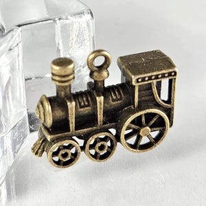 May include: A miniature bronze-colored train engine with detailed wheels, smokestack, and cabin. The vintage-style locomotive is a decorative item, possibly a charm or a small figurine, with an antique finish.