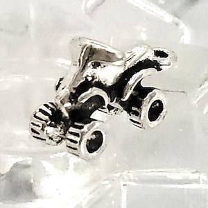 May include: A silver charm in the shape of a monster truck. The truck has four wheels and a detailed design.