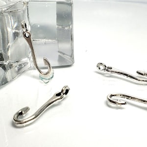 Puede incluir: Tres dijes de metal plateado con forma de anzuelo, cada uno con un vástago curvo y un pequeño lazo circular en la parte superior para su fijación. Los dijes tienen un acabado pulido y reflectante y están dispuestos en grupo sobre una superficie clara.