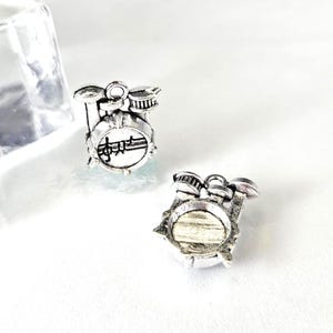 May include: Two silver-colored drum set charms. Each charm features a miniature drum set with a bass drum, snare drum, and cymbals. The bass drum has musical notes on it. The charms are on a white surface.